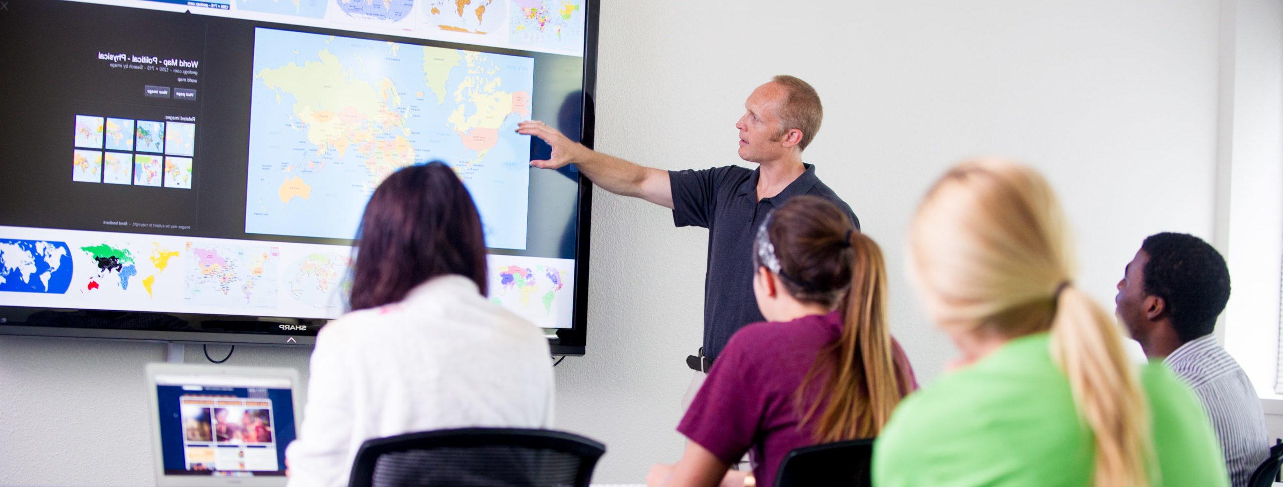 Man teaching students pointing to projector screen displaying world maps.