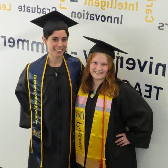 Two students, each dressed in a cap and gown, pose against a word-cloud backdrop.
